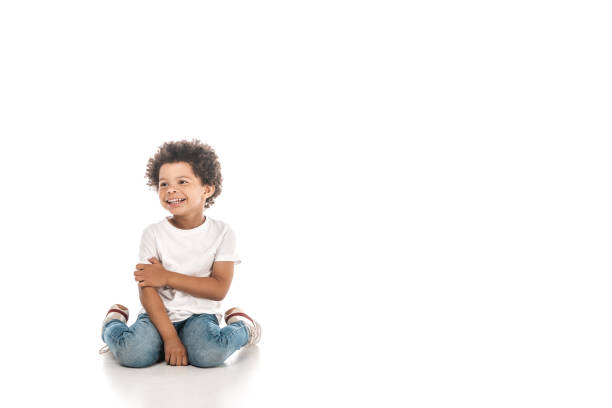 alegre niño afroamericano mirando hacia otro lado mientras se sienta sobre fondo blanco - sentado en el suelo fotografías e imágenes de stock