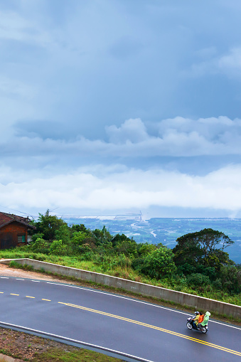 Khmer family driving motorcycle on the mountain asphalt road on a rainy day, cloudy covered the Gulf in the background. Kampot, South Cambodia.