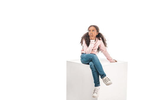 thoughtful african american child looking away while sitting on white cube isolated on white