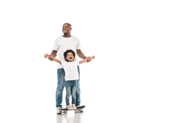 Photo of happy african american man supporting cheerful son standing on penny board on white background