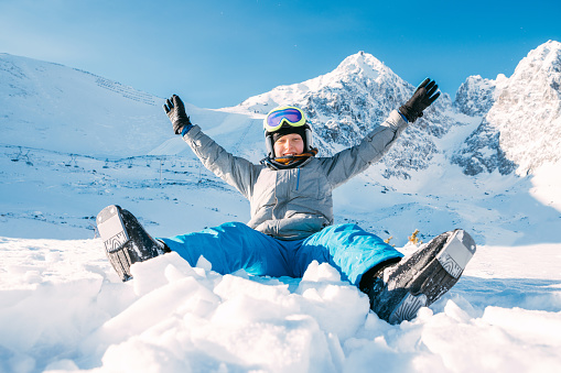 Portrait of laughing boy sitting on the snowy slope, rising an arms up with High Tatra Mountains range background. Carefree childhood winter holidays concept image.