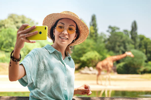 счастливая азиатская зоология студентка делает селфи фото на смартфоне, пока жираф пьет из озера - people adventure vacations tropical climate стоковые фото и изображения
