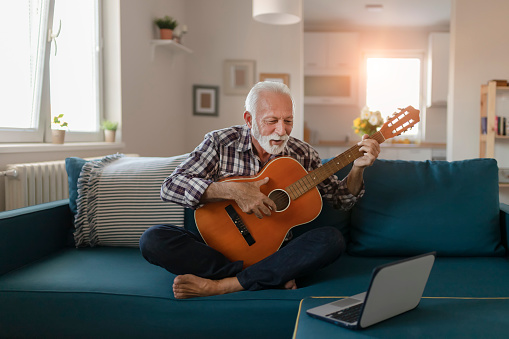 Happy good looking elderly man in a plaid shirt sits on a sofa in the living room and learns to play acoustic guitar online using a Laptop