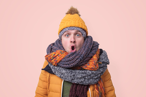 Handsome caucasian shocked funny man in several hats and scarfs being surprised on pink background. Winter fashion.