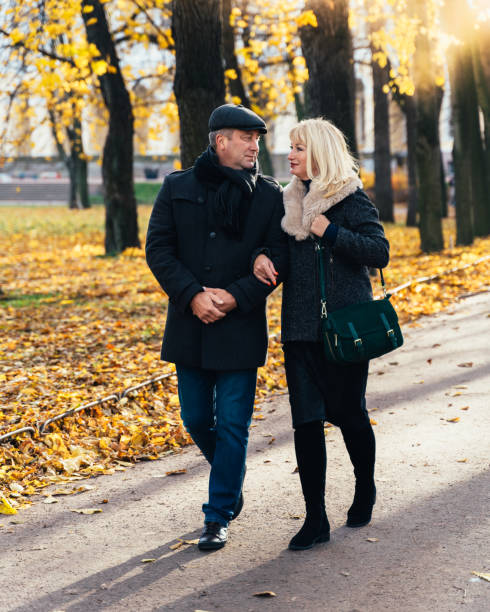 felice donna bionda matura e bell'uomo bruna di mezza età camminano nel parco, guardandoti l'un l'altro. coppia amorevole di passeggiate di 45-50 anni nel parco autunnale in abiti caldi - 40 45 years immagine foto e immagini stock