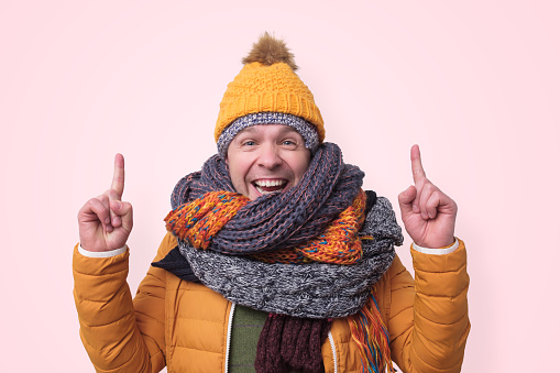 Handsome caucasian funny man in several hats and scarfs pointing with index finger up on pink background. Winter fashion.
