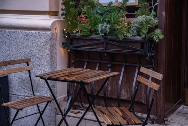 istock Wooden chairs and table near window decorated with green conifer tree branches, red berries and shiny golden Christmas lights with old stone wall background. Winter holiday decorations. New Year scene 1187155043