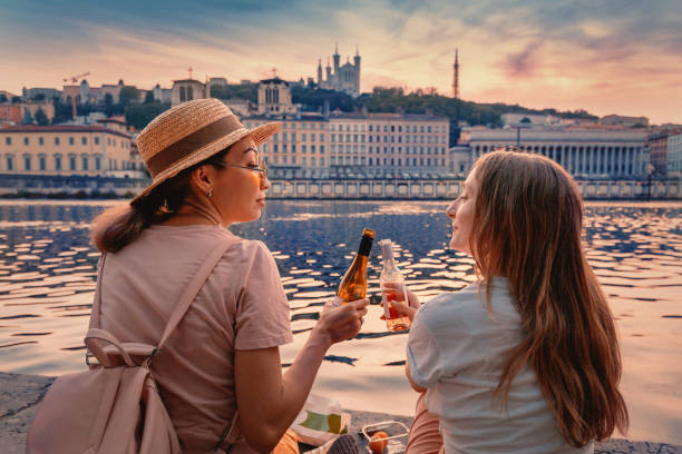 two happy girls close friends celebrating and toasting and drinking wine from a little bottles and watching a sun goes down over saone river in lyon city, france - drinking little girls women wine imagens e fotografias de stock