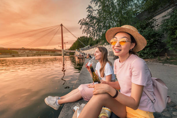 two happy girls close friends celebrating and toasting and drinking wine from a little bottles and watching a sun goes down over saone river in lyon city, france - drinking little girls women wine imagens e fotografias de stock