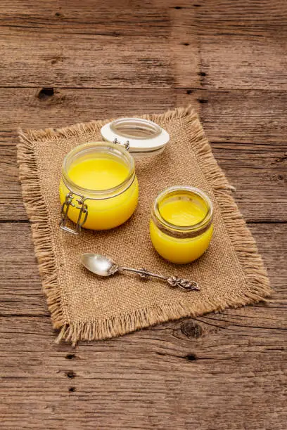 Photo of Pure or desi ghee (ghi), clarified melted butter. Healthy fats bulletproof diet concept or paleo style plan. Glass jars, silver spoon on vintage sackcloth. Wooden boards background