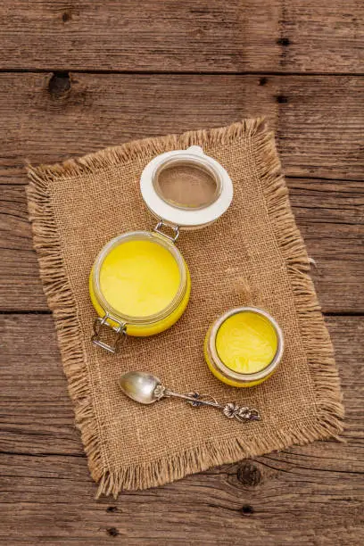 Photo of Pure or desi ghee (ghi), clarified melted butter. Healthy fats bulletproof diet concept or paleo style plan. Glass jars, silver spoon on vintage sackcloth. Wooden boards background