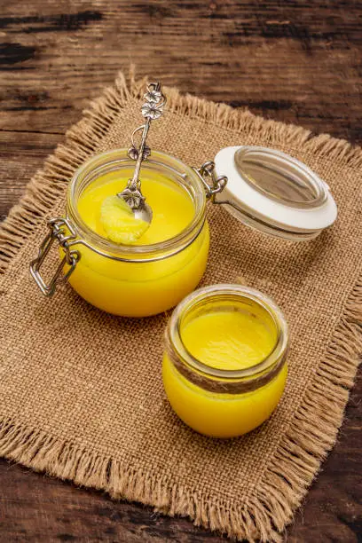 Photo of Pure or desi ghee (ghi), clarified melted butter. Healthy fats bulletproof diet concept or paleo style plan. Glass jars, silver spoon on vintage sackcloth. Wooden boards background