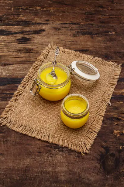 Photo of Pure or desi ghee (ghi), clarified melted butter. Healthy fats bulletproof diet concept or paleo style plan. Glass jars, silver spoon on vintage sackcloth. Wooden boards background