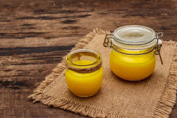 Photo of Pure or desi ghee (ghi), clarified melted butter. Healthy fats bulletproof diet concept or paleo style plan. Glass jars, silver spoon on vintage sackcloth. Wooden boards background