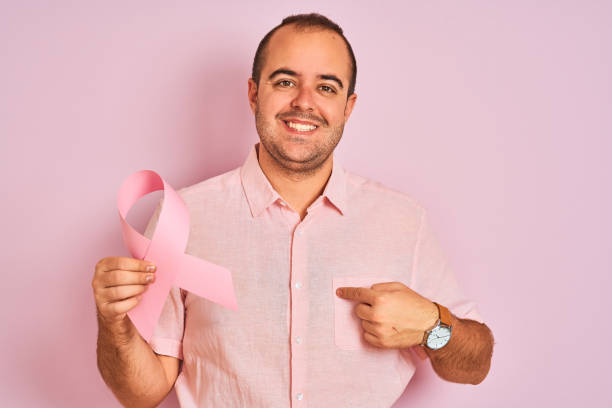 Young man holding cancer ribbon symbol standing over isolated pink background with surprise face pointing finger to himself Young man holding cancer ribbon symbol standing over isolated pink background with surprise face pointing finger to himself brest cancer hope stock pictures, royalty-free photos & images