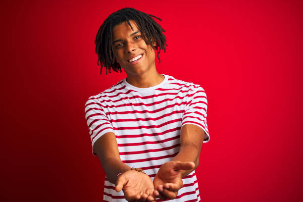 giovane afro uomo con dreadlocks che indossa t-shirt a righe in piedi su uno sfondo rosso isolato sorridente con le mani palmi delle mani insieme ricevendo o dando gesto. attesa e protezione - human hand palm reaching fashion model foto e immagini stock