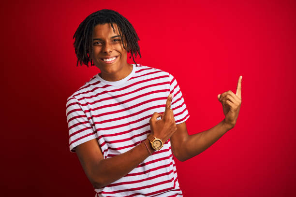 istock Young afro man with dreadlocks wearing striped t-shirt standing over isolated red background smiling and looking at the camera pointing with two hands and fingers to the side. 1187190831