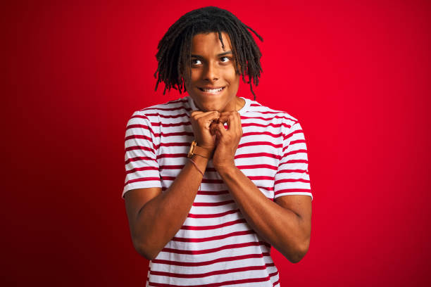 istock Young afro man with dreadlocks wearing striped t-shirt standing over isolated red background laughing nervous and excited with hands on chin looking to the side 1187190874