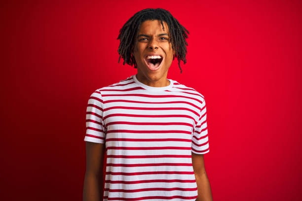 istock Young afro man with dreadlocks wearing striped t-shirt standing over isolated red background angry and mad screaming frustrated and furious, shouting with anger. Rage and aggressive concept. 1187190882
