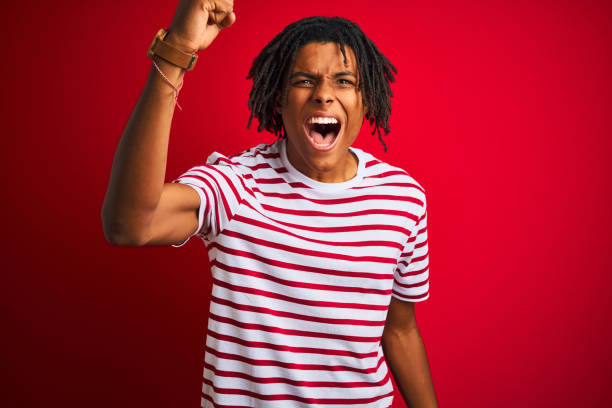 istock Young afro man with dreadlocks wearing striped t-shirt standing over isolated red background angry and mad raising fist frustrated and furious while shouting with anger. Rage and aggressive concept. 1187190896