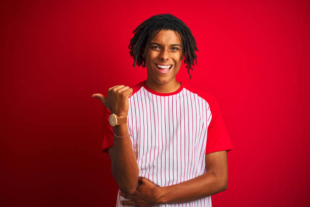 istock Afro man with dreadlocks wearing striped t-shirt standing over isolated red background smiling with happy face looking and pointing to the side with thumb up. 1187190924