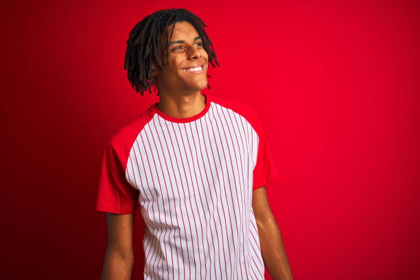 istock Afro man with dreadlocks wearing striped t-shirt standing over isolated red background looking away to side with smile on face, natural expression. Laughing confident. 1187190956