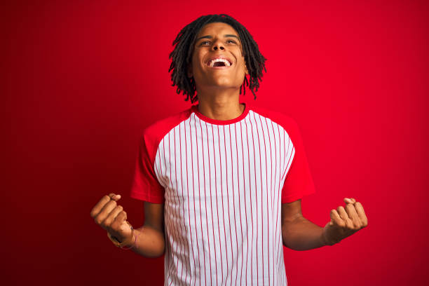 istock Afro man with dreadlocks wearing striped t-shirt standing over isolated red background celebrating surprised and amazed for success with arms raised and open eyes. Winner concept. 1187190963