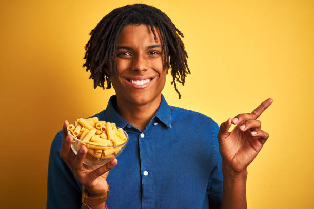 istock Afro american man with dreadlocks holding pasta macaroni over isolated yellow background very happy pointing with hand and finger to the side 1187191468