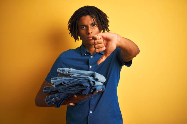 homme d'afro avec des dreadlocks retenant la pile des jeans pliés au-dessus du fond jaune d'isolement avec le visage fâché, signe négatif affichant l'aversion avec des pouces vers le bas, concept de rejet - washing hair photos et images de collection