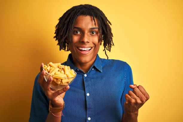 istock Afro american man with dreadlocks holding pasta macaroni over isolated yellow background screaming proud and celebrating victory and success very excited, cheering emotion 1187191500