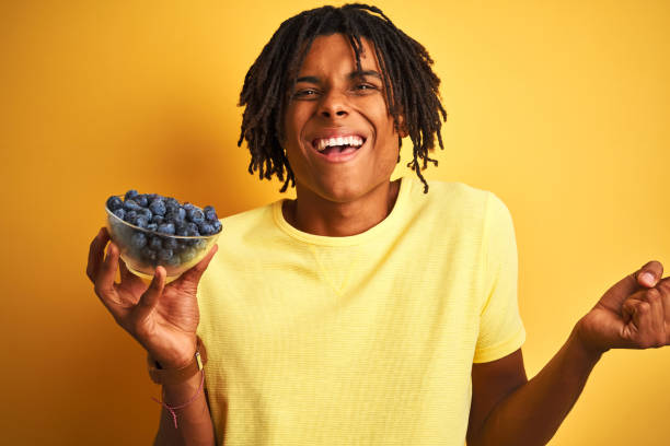istock Afro american man with dreadlocks holding blueberries over isolated yellow background screaming proud and celebrating victory and success very excited, cheering emotion 1187193228