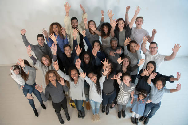 A large group of ethnically diverse individuals of varying ages and ethnicity's, stand for a portrait as they raise their hands above their heads.  They are all wearing casual clothing and smiling.  The photo is an aerial view.