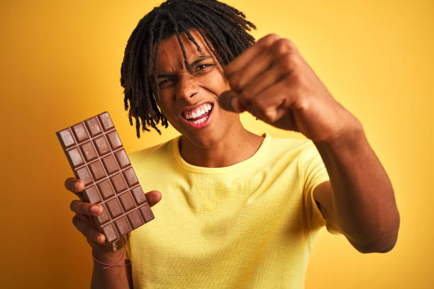 istock Afro american man with dreadlocks eating chocolate bar over isolated yellow background annoyed and frustrated shouting with anger, crazy and yelling with raised hand, anger concept 1187193358