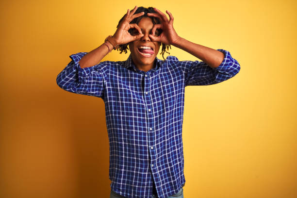 istock Afro man with dreadlocks wearing casual shirt standing over isolated yellow background doing ok gesture like binoculars sticking tongue out, eyes looking through fingers. Crazy expression. 1187193427