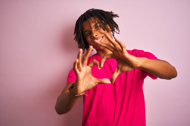 homem americano afro com dreadlocks vestindo t-shirt de pé sobre fundo rosa isolado sorrindo no amor mostrando símbolo do coração e forma com as mãos. conceito romântico. - jovem no coração - fotografias e filmes do acervo