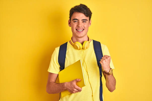 adolescente ragazzo che indossa cuffie e zaino leggendo un libro su sfondo isolato urlando orgoglioso e celebrando vittoria e successo molto eccitato, tifo emozione - university freshman portrait teenager foto e immagini stock