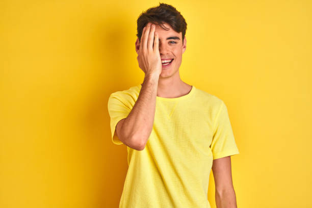 menino adolescente que desgasta o t-shirt amarelo sobre o fundo isolado que cobre um olho com mão, sorriso confiável na cara e emoção da surpresa. - half smile - fotografias e filmes do acervo