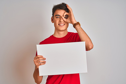 Teenager boy holding advertising banner with blank space over isolated background with happy face smiling doing ok sign with hand on eye looking through fingers