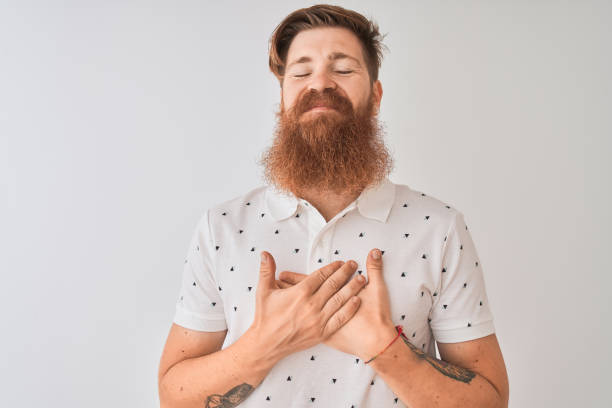Young redhead irish man wearing polo standing over isolated white background smiling with hands on chest with closed eyes and grateful gesture on face. Health concept. Young redhead irish man wearing polo standing over isolated white background smiling with hands on chest with closed eyes and grateful gesture on face. Health concept. chest tattoos for men designs stock pictures, royalty-free photos & images