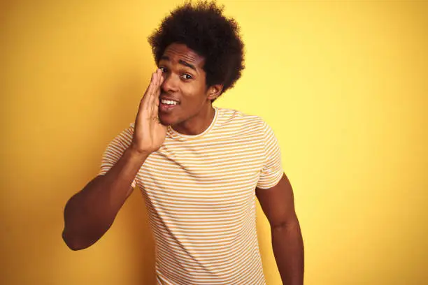 American man with afro hair wearing striped t-shirt standing over isolated yellow background hand on mouth telling secret rumor, whispering malicious talk conversation