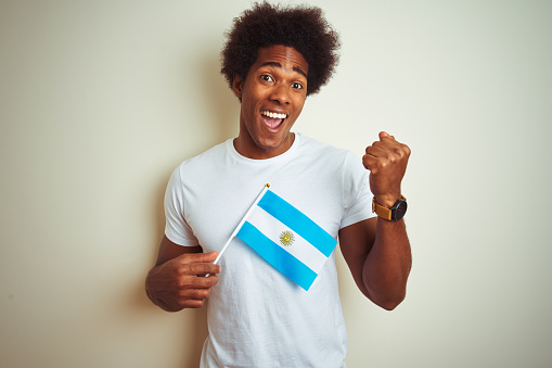 Afro american tourist man holding Argentinian Argentina flag over isolated white background screaming proud and celebrating victory and success very excited, cheering emotion