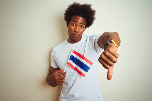 Young afro american man holding Thailand Thai flag standing over isolated white background with angry face, negative sign showing dislike with thumbs down, rejection concept