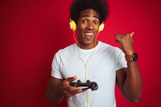 istock Afro gamer man playing video game using joystick headphones over isolated red background pointing and showing with thumb up to the side with happy face smiling 1187198727