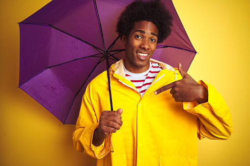 Afro american man wearing rain coat and umbrella standing over isolated yellow background with surprise face pointing finger to himself