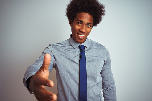 American business man with afro hair wearing shirt and tie over isolated white background smiling friendly offering handshake as greeting and welcoming. Successful business.