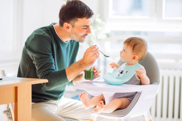Hungry baby Father feeding baby boy with spoon in high chair, they enjoy in togetherness high chair stock pictures, royalty-free photos & images