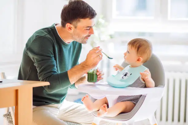 Father feeding baby boy with spoon in high chair, they enjoy in togetherness