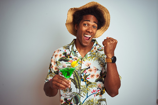 Afro man on holidays wearing summer hat drinking cocktail over isolated white background screaming proud and celebrating victory and success very excited, cheering emotion