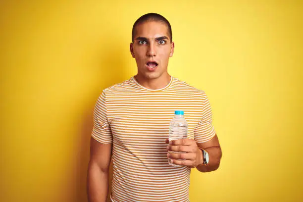 Photo of Young handsome man holding plastic water bottle over yellow isolated background scared in shock with a surprise face, afraid and excited with fear expression