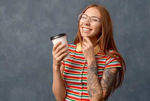 Thoughtful woman holds chin, enjoys drinking takeaway coffee, has natural beauty. Beautiful girl ginger long hair and tattoo on arm, wearing striped top big round glasses, isolated on wall in Studio